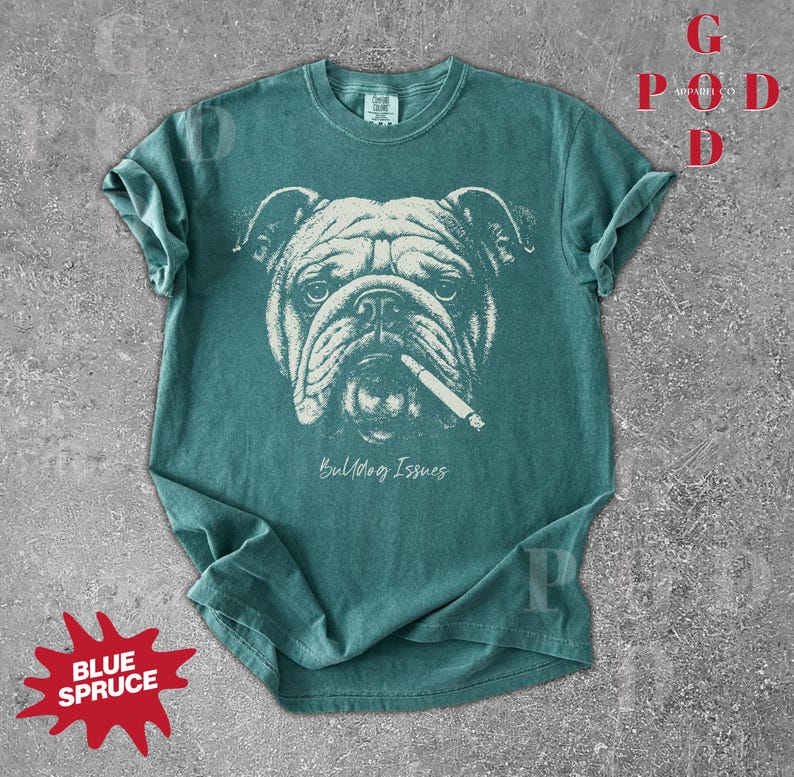 May include: Teal-colored t-shirt featuring a detailed graphic of a bulldog with a cigarette in its mouth. The text "Bulldog Issues" is printed below the image. The shirt has rolled-up sleeves and a crew neck.