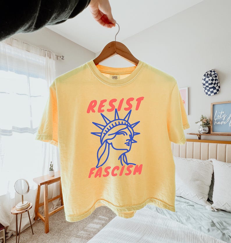 May include: Yellow t-shirt with the words "RESIST FASCISM" in red and a blue graphic of the Statue of Liberty. The shirt is hanging on a wooden hanger.