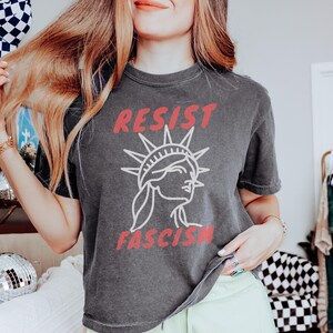 May include: Dark gray t-shirt with the words "RESIST FASCISM" in red and a white illustration of the Statue of Liberty. The shirt is a casual style, suitable for everyday wear.