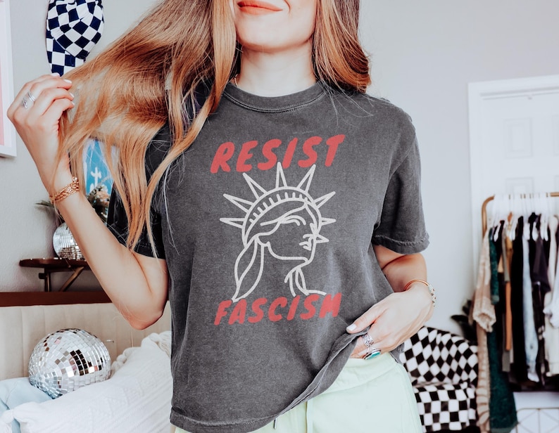 May include: Dark gray t-shirt with the words "RESIST FASCISM" in red and a white illustration of the Statue of Liberty. The shirt is a casual style, suitable for everyday wear.