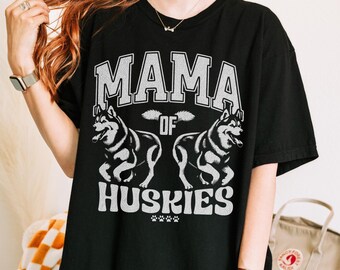May include: Black t-shirt with the text "MAMA of Huskies" in a white, distressed font. Two husky dog illustrations are featured below the text. Paw prints are at the bottom of the design.