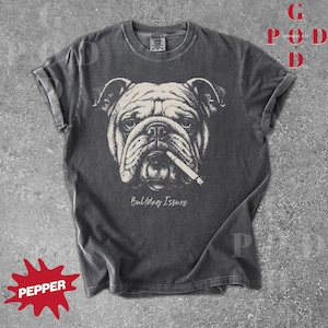 May include: Dark gray t-shirt featuring a detailed illustration of a bulldog with a cigarette in its mouth. The graphic is in black and white, with the words "Bulldog Issues" below the image. The shirt has rolled-up sleeves.