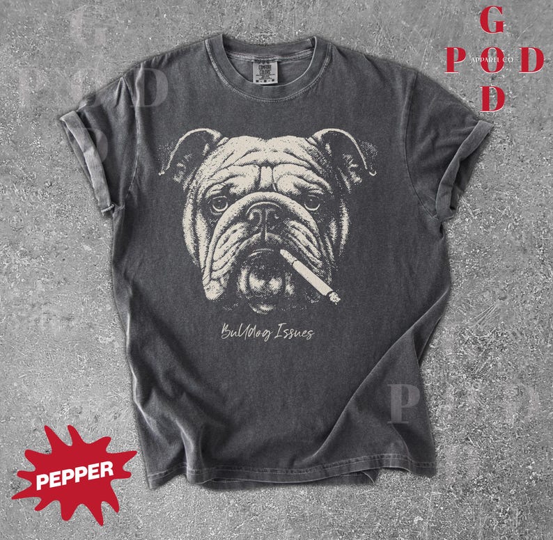 May include: Dark gray t-shirt featuring a detailed illustration of a bulldog with a cigarette in its mouth. The graphic is in black and white, with the words "Bulldog Issues" below the image. The shirt has rolled-up sleeves.