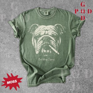 May include: A moss green t-shirt featuring a detailed illustration of a bulldog with a cigarette in its mouth. The text "Bulldog Issues" is printed below the image. The shirt has rolled-up sleeves.