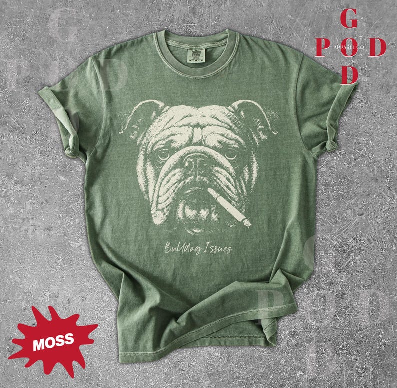 May include: A moss green t-shirt featuring a detailed illustration of a bulldog with a cigarette in its mouth. The text "Bulldog Issues" is printed below the image. The shirt has rolled-up sleeves.