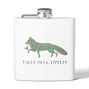Puede incluir: Una petaca de metal blanco con tapón de rosca plateado. El frente presenta una ilustración de un zorro verde azulado estilizado con patrones florales naranjas, sosteniendo una copa de martini. Debajo, el texto TALLY HO & TIPPLES en gris oscuro.