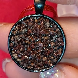 Pode incluir: Um pingente redondo com uma moldura de metal escuro preenchida com pequenas pedras marrons e vermelho escuro. O pingente pende de um fino cordão vermelho. O fundo é de cor vermelha sólida.