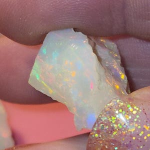 May include: A raw opal gemstone, held between fingers. The stone has a milky white base colour and displays iridescent flashes of green, yellow, orange, and pink. The opal has a rough, natural texture.