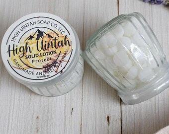 Könnte beinhalten: Ein Glas mit fester Lotion und ein Glas mit weißen, runden Lotiontabletten. Das Glas mit fester Lotion trägt die Aufschrift "High Uintah SOLID LOTION Protect HANDMADE AND ALL NATURAL 62 gr". Ein Holzlöffel und ein Lavendelzweig sind ebenfalls zu sehen.