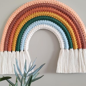 Könnte beinhalten: Ein handgefertigter Makramee-Regenbogen zum Aufhängen an der Wand. Der Regenbogen hat Farben wie Pfirsich, Braun, Orange, Gelb, Grün, Blau und Weiß. Am unteren Ende befinden sich weiße Fransen. Ein dekorativer Artikel.
