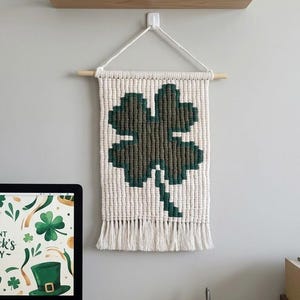 Può includere: Un arazzo in macramè con un motivo a trifoglio a quattro foglie verde e verde scuro. L'arazzo rettangolare ha una frangia nella parte inferiore ed è sospeso a un tassello di legno. Una decorazione per il giorno di San Patrizio.