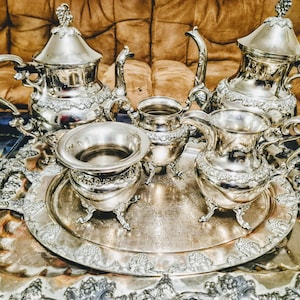May include: An antique silver tea set comprising a teapot, milk jug, sugar bowl, and serving tray. The set displays elaborate floral patterns and curved handles. The pieces are arranged on a decorative tray, showcasing a classic design.