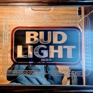 Puede incluir: Espejo enmarcado con el logotipo de Bud Light. El logotipo rectangular tiene un borde rojo y letras doradas "BUD LIGHT". La palabra "BEER" está debajo. El marco es marrón oscuro, con detalles de trigo y águila.