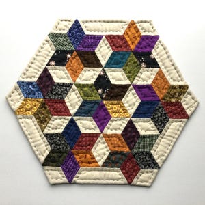 May include: A hexagonal quilted mat featuring a geometric pattern of multicolored fabric diamonds in shades of purple, orange, blue, green, red, and black, separated by cream-colored fabric strips with visible white hand-stitched quilting lines.