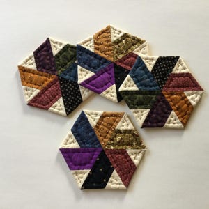 May include: A collection of six-sided fabric coasters. Each coaster is made of various patterned fabrics in shades of brown, blue, purple, and green, with a cream-colored border. The coasters are arranged on a white surface.