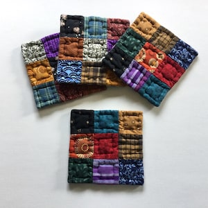 May include: Three square fabric coasters featuring a patchwork design of nine smaller quilted squares. Each coaster displays a variety of colors and patterns, including floral, plaid, and geometric prints in shades of red, blue, green, and brown.