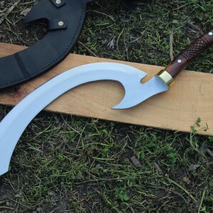 May include: A curved, silver-colored sword with a wooden handle and brass accents. The sword is resting on a wooden surface.