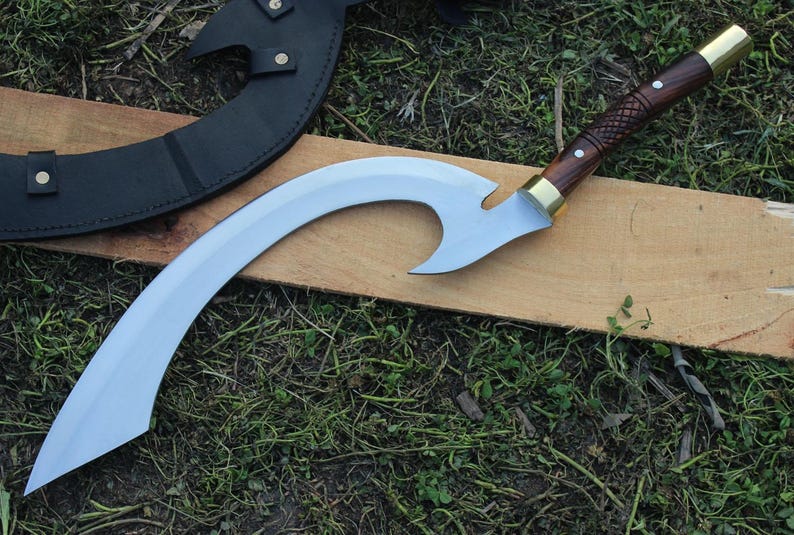 May include: A curved, silver-colored sword with a wooden handle and brass accents. The sword is resting on a wooden surface.