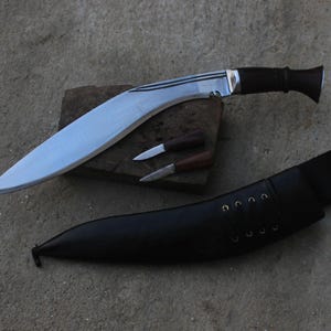 May include: A large, curved blade knife with a dark wooden handle and a smaller knife set. The blade is silver and the handle is black. A black leather sheath with eyelets is also included. The knives are displayed on a wooden block.