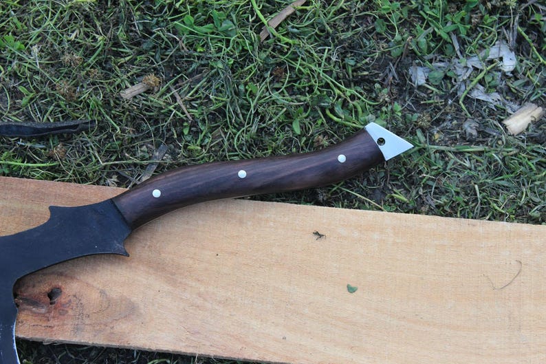 May include: A black axe with a dark brown wooden handle and silver accents. The axe is lying on a wooden surface.