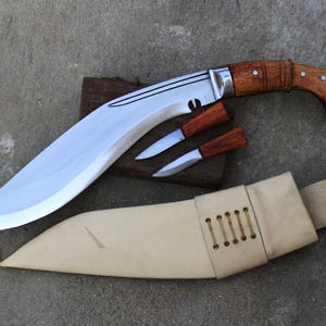 May include: A large, curved blade knife with a wooden handle and two smaller knives, all with wooden handles. The knives are displayed with a tan leather sheath. The blades are silver and the handles are a light brown colour.