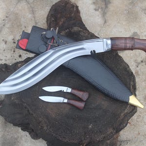 May include: A large, curved blade knife with a wooden handle and a smaller knife, both with sheaths. The blade has a silver finish with a wave pattern. The handle is dark brown. The sheath is black with a gold-colored tip.