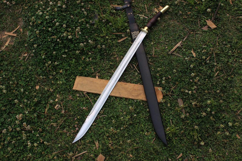 Pu&ograve; includere: Una spada argentata con un manico in legno e un fodero in pelle nera. La spada &egrave; appoggiata su una superficie in legno e uno sfondo di erba verde.