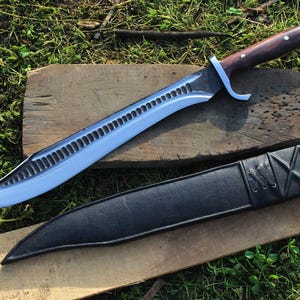 May include: A silver and brown knife with a serrated blade and a black leather sheath. The knife is lying on a wooden surface with green grass in the background.