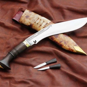 May include: A large, curved blade knife with a wooden handle and a decorative metal bolster. The knife rests on a brown leather surface, accompanied by a sheath and two smaller knives. The blade is silver, and the handle is dark brown.