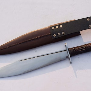 May include: A large, silver-bladed knife with a dark brown, carved handle and a brown leather sheath. The sheath has a black leather belt loop with rivets. The knife is designed for utility and display.