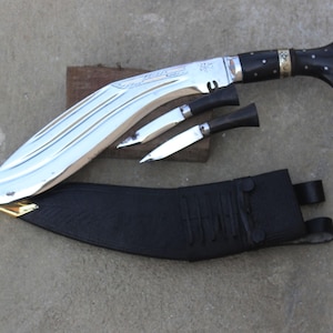 Könnte beinhalten: Ein großes Messer mit gebogener Klinge, polierter Silberoberfläche, schwarzem Griff und zwei kleineren Messern. Das Set enthält eine schwarze Lederscheide. Das Messer wird auf einem Holzblock präsentiert.