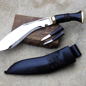May include: A large, curved blade knife with a polished steel finish, black handle, and gold accents. Two smaller knives and a black leather sheath are also present. The knife rests on a wooden block.