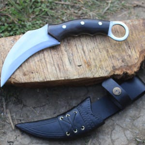 Puede incluir: Un cuchillo karambit curvo con mango negro y hoja plateada, apoyado sobre una superficie de madera. El cuchillo va acompa&ntilde;ado de una funda de cuero negro con costuras decorativas y cierre de bot&oacute;n a presi&oacute;n.