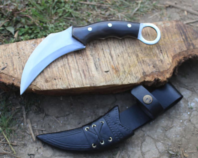 Puede incluir: Un cuchillo karambit curvo con mango negro y hoja plateada, apoyado sobre una superficie de madera. El cuchillo va acompa&ntilde;ado de una funda de cuero negro con costuras decorativas y cierre de bot&oacute;n a presi&oacute;n.