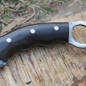 Puede incluir: Primer plano de un cuchillo curvo con mango negro y hoja plateada. El mango tiene tres incrustaciones de puntos blancos. El cuchillo tiene un anillo para el dedo al final del mango. El cuchillo est&aacute; sobre una superficie de madera.