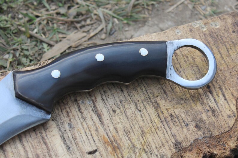 Puede incluir: Primer plano de un cuchillo curvo con mango negro y hoja plateada. El mango tiene tres incrustaciones de puntos blancos. El cuchillo tiene un anillo para el dedo al final del mango. El cuchillo est&aacute; sobre una superficie de madera.