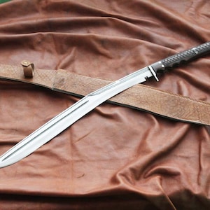 Puede incluir: Una espada de hoja plateada con un mango oscuro y una funda de cuero marrón. La espada está en ángulo sobre la funda, apoyada sobre una superficie de tela marrón. El mango tiene un diseño texturizado.