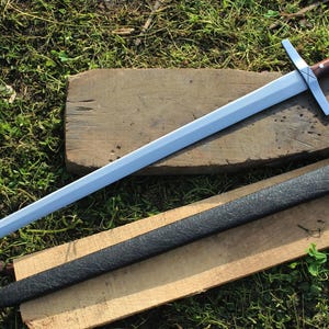 May include: A silver sword with a brown wooden handle and a black leather scabbard. The sword is lying on a wooden surface with a green grassy background.
