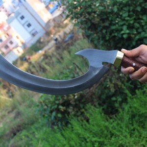 May include: A curved, black metal blade with a wooden handle and brass accents. The blade is held by a hand in front of a blurred background.