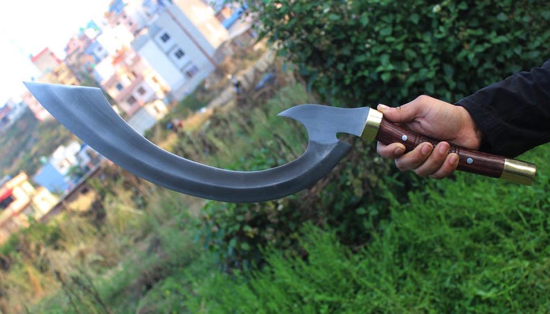 May include: A curved, black metal blade with a wooden handle and brass accents. The blade is held by a hand in front of a blurred background.