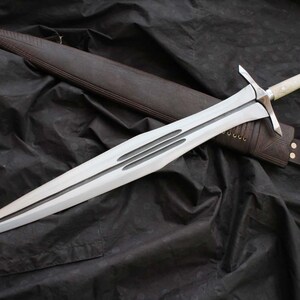 Puede incluir: Una espada plateada con una hoja de doble filo y una guarda, sobre una superficie negra. La espada tiene un mango de color claro y va acompañada de una funda de cuero marrón.