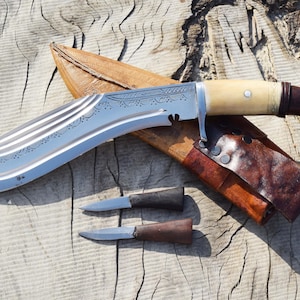 May include: A large, curved kukri knife with a silver blade and a bone and dark wood handle, resting on a wooden surface. Includes a leather sheath and two smaller knives. The blade has decorative etchings.