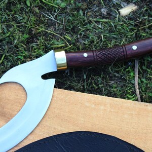 May include: A curved, silver-colored blade with a wooden handle and brass accents. The handle is decorated with a carved pattern and has a white dot on the top.