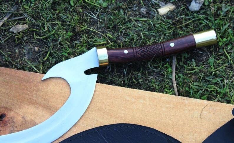 May include: A curved, silver-colored blade with a wooden handle and brass accents. The handle is decorated with a carved pattern and has a white dot on the top.
