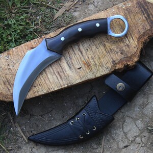 Puede incluir: Un cuchillo karambit curvo con mango negro y hoja plateada, sobre una superficie de madera. El cuchillo tiene un anillo para el dedo y va acompa&ntilde;ado de una funda de cuero negro.