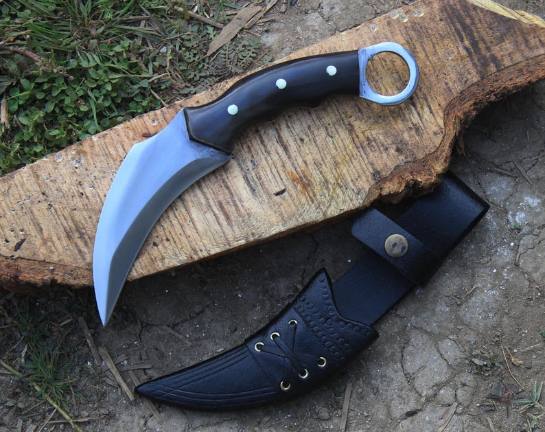 Puede incluir: Un cuchillo karambit curvo con mango negro y hoja plateada, sobre una superficie de madera. El cuchillo tiene un anillo para el dedo y va acompa&ntilde;ado de una funda de cuero negro.