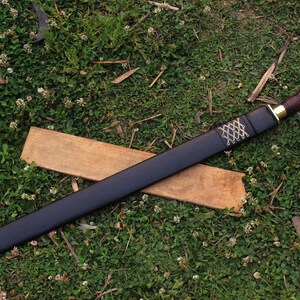 Pu&ograve; includere: Un fodero in pelle nera con una punta in ottone e un motivo intrecciato decorativo. Il fodero &egrave; appoggiato su una superficie di legno e erba verde.