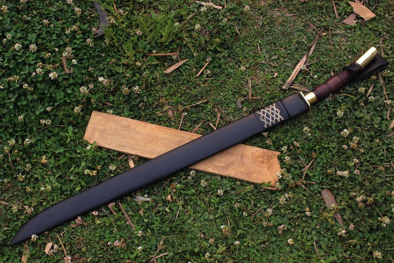 Pu&ograve; includere: Un fodero in pelle nera con una punta in ottone e un motivo intrecciato decorativo. Il fodero &egrave; appoggiato su una superficie di legno e erba verde.