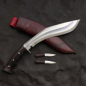 May include: A large, curved blade knife with a dark wooden handle and a polished silver blade. The knife rests on a dark surface, accompanied by a leather sheath and two smaller knives with wooden handles. The sheath is brown with a red interior.
