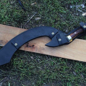 May include: A black leather scabbard with a curved blade and a wooden handle. The scabbard has a brass button closure and a decorative design.